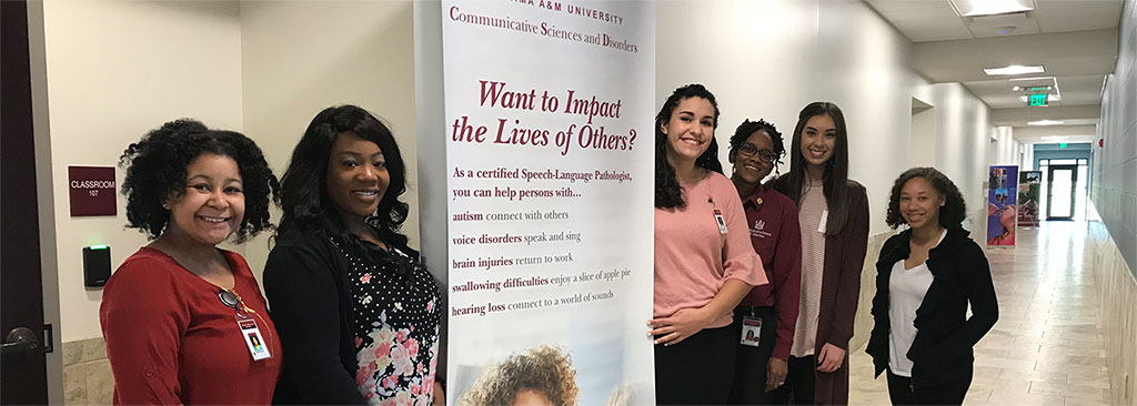 Smiling students pose next to a promotional poster for the Communicative Sciences and Disorders program