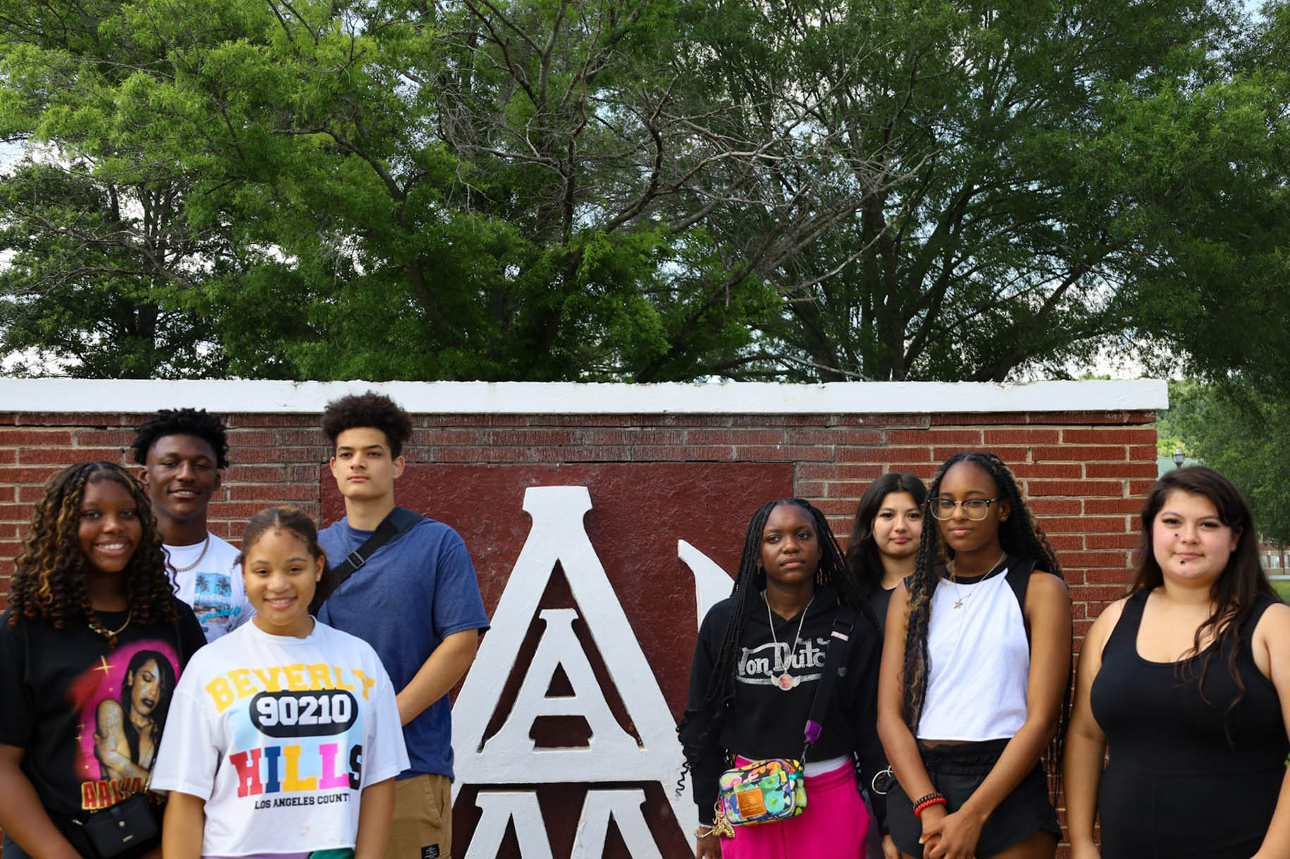 Two male high school students and six female high school students stand in front of the AAMU Interlock logo at the main gate of campus