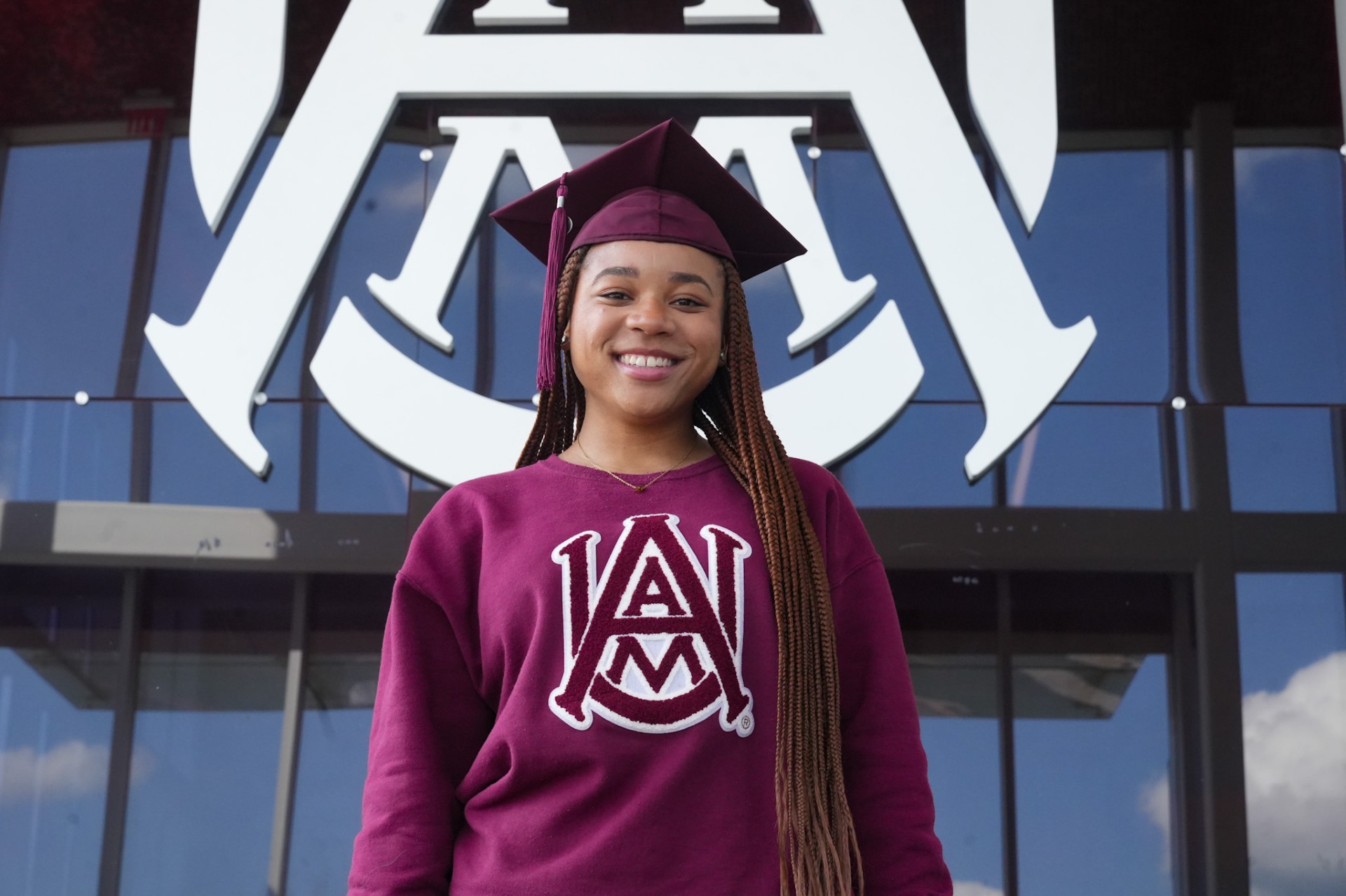 Amirah Ali wears sweater with AAMU interlock logo and maroon graduation cap in front of the Event Center