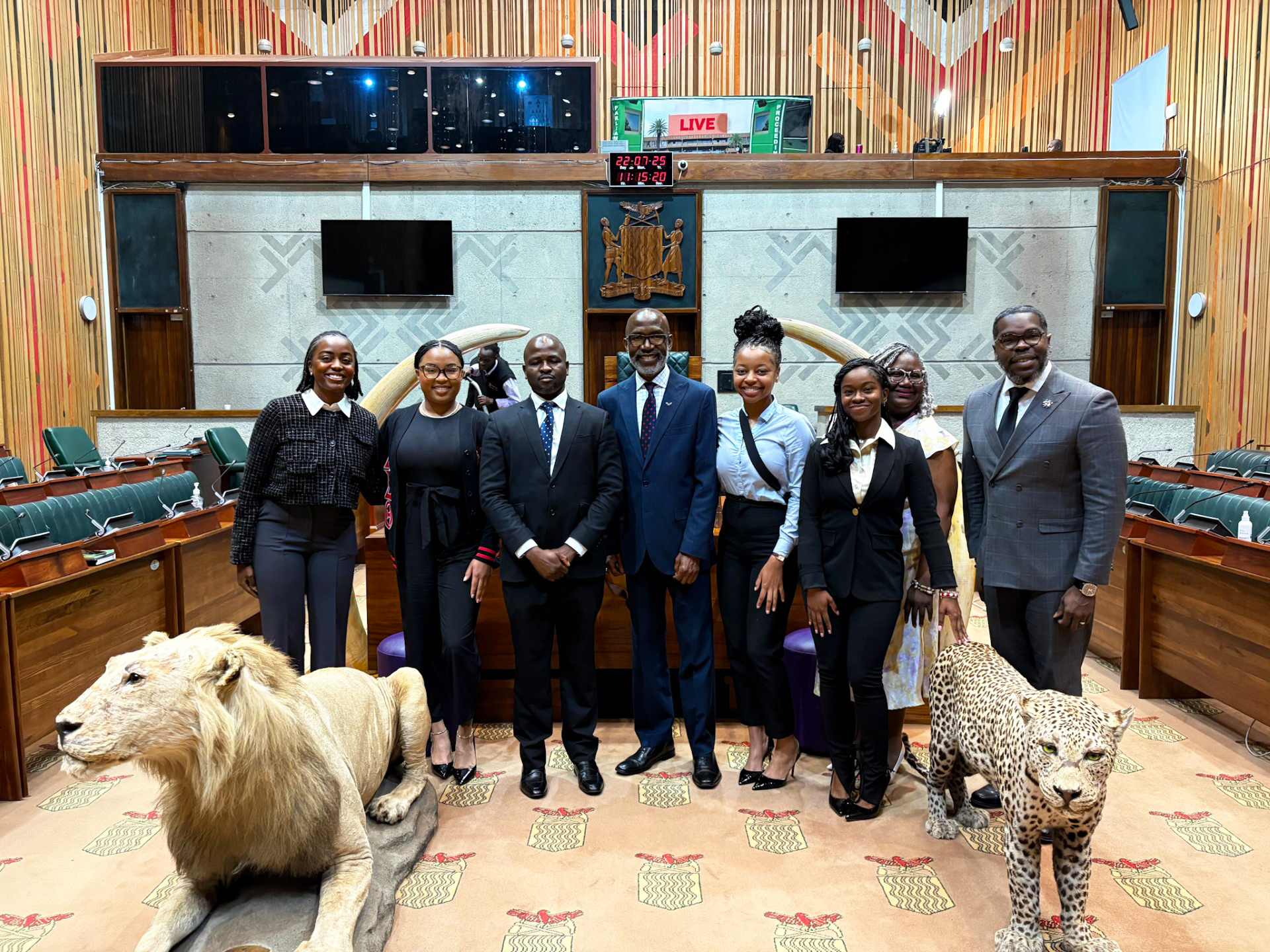 ) Michayla Johnson, Desiree' Munford, Zambia National Assembly official, Dr. Samson Chama, Angel Hayes, Nyla Lofton, Jacobie Kelsey, Professor Willie Diggs post with a stuffed lion and tiger in a school room in Zambia