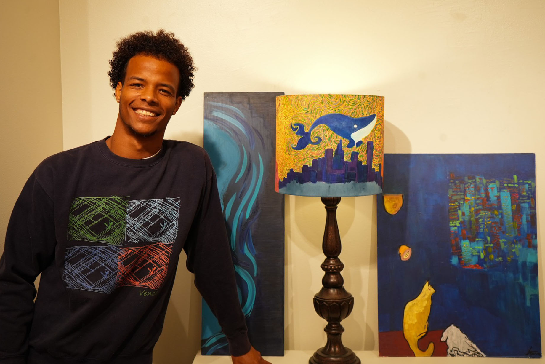 Vence N'tcha stands next to his paintings of his lamp and his cats