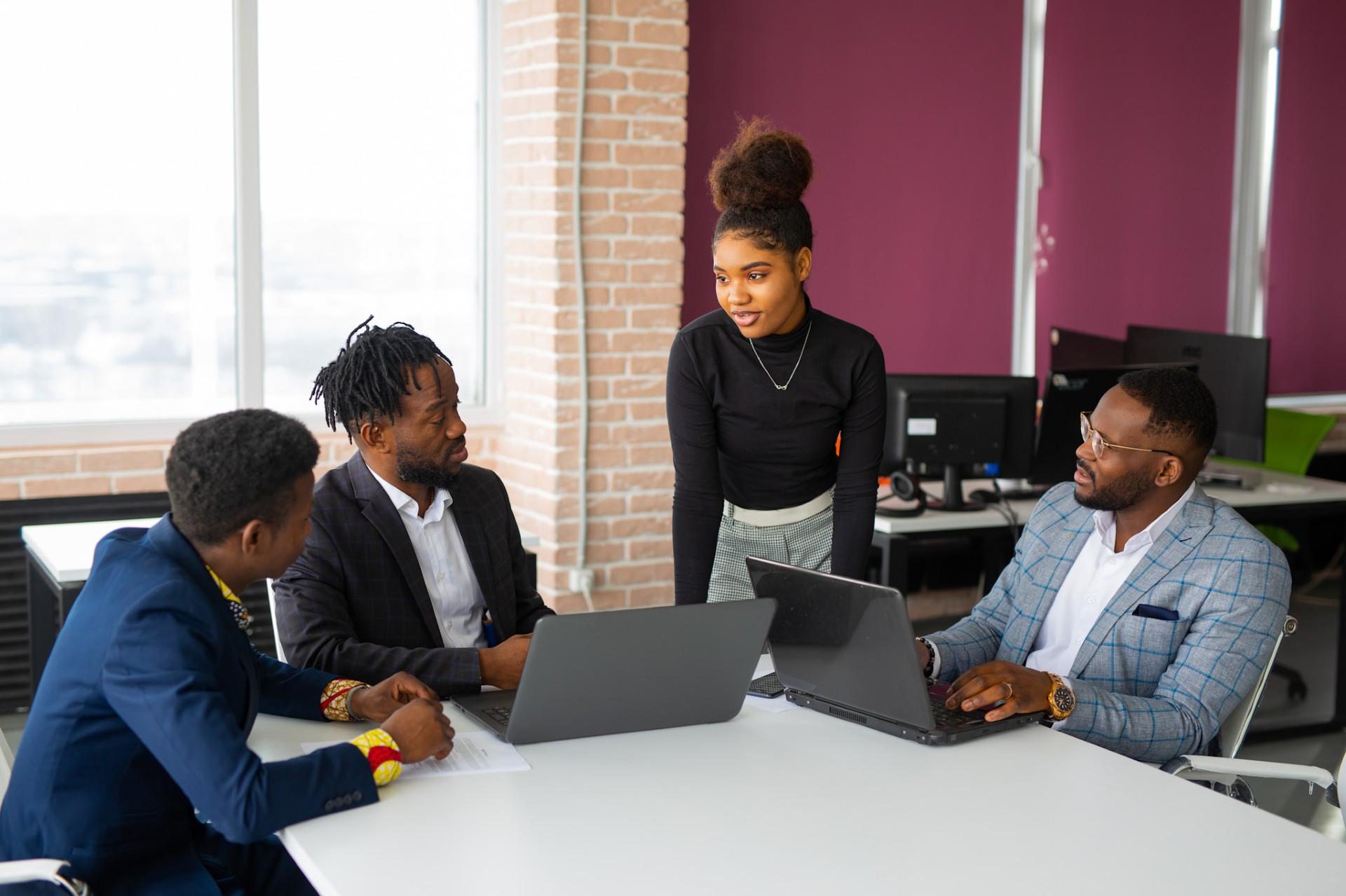 Female employee stands at table talking to three seated male colleagues, two using computers