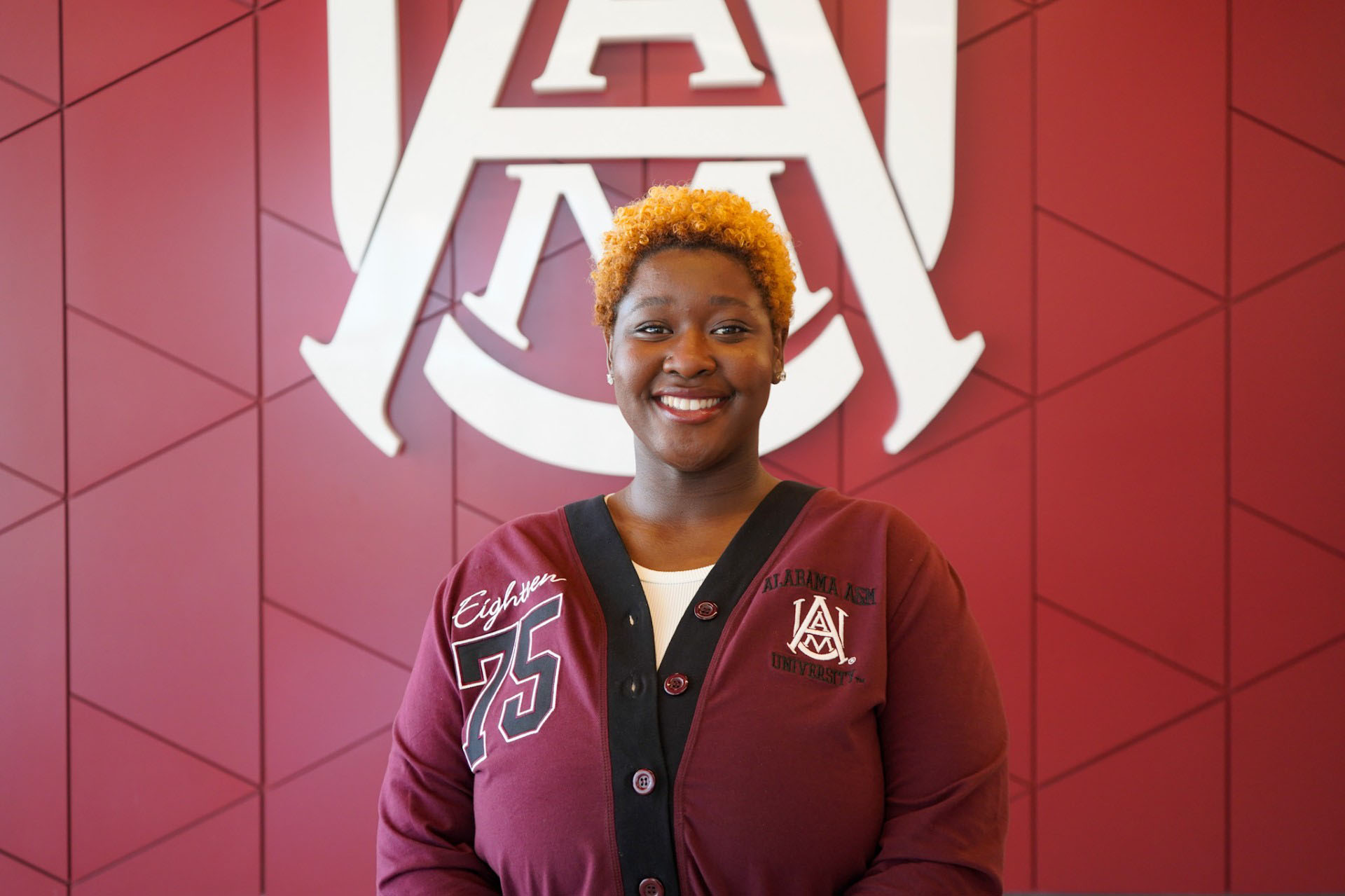 Taylor Gamble smiles and wears an Alabama A&M sweater in front of the interlock logo in the lobby of the Event Center