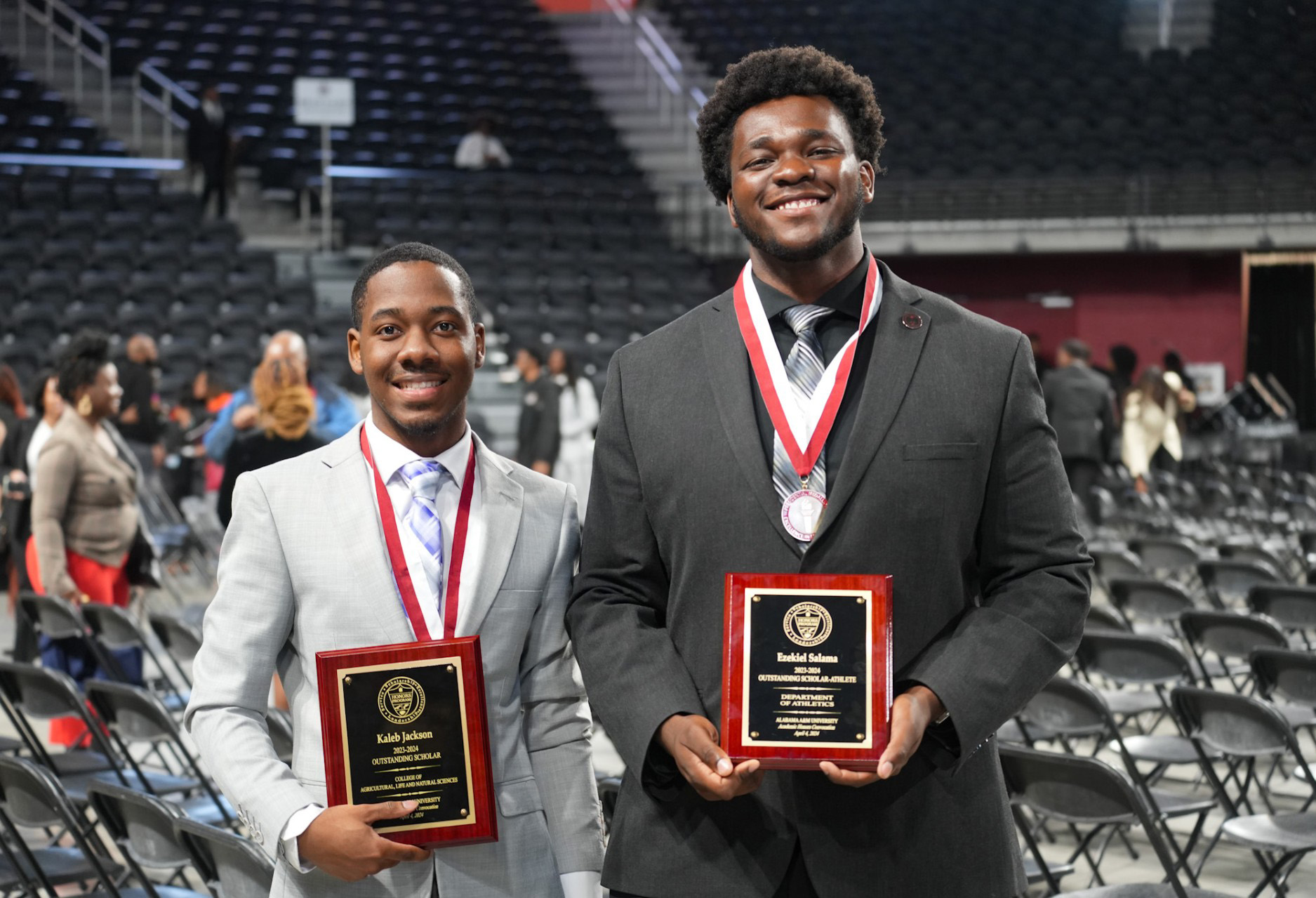 Two male students smile while wearing academic medallions and holding academic awards