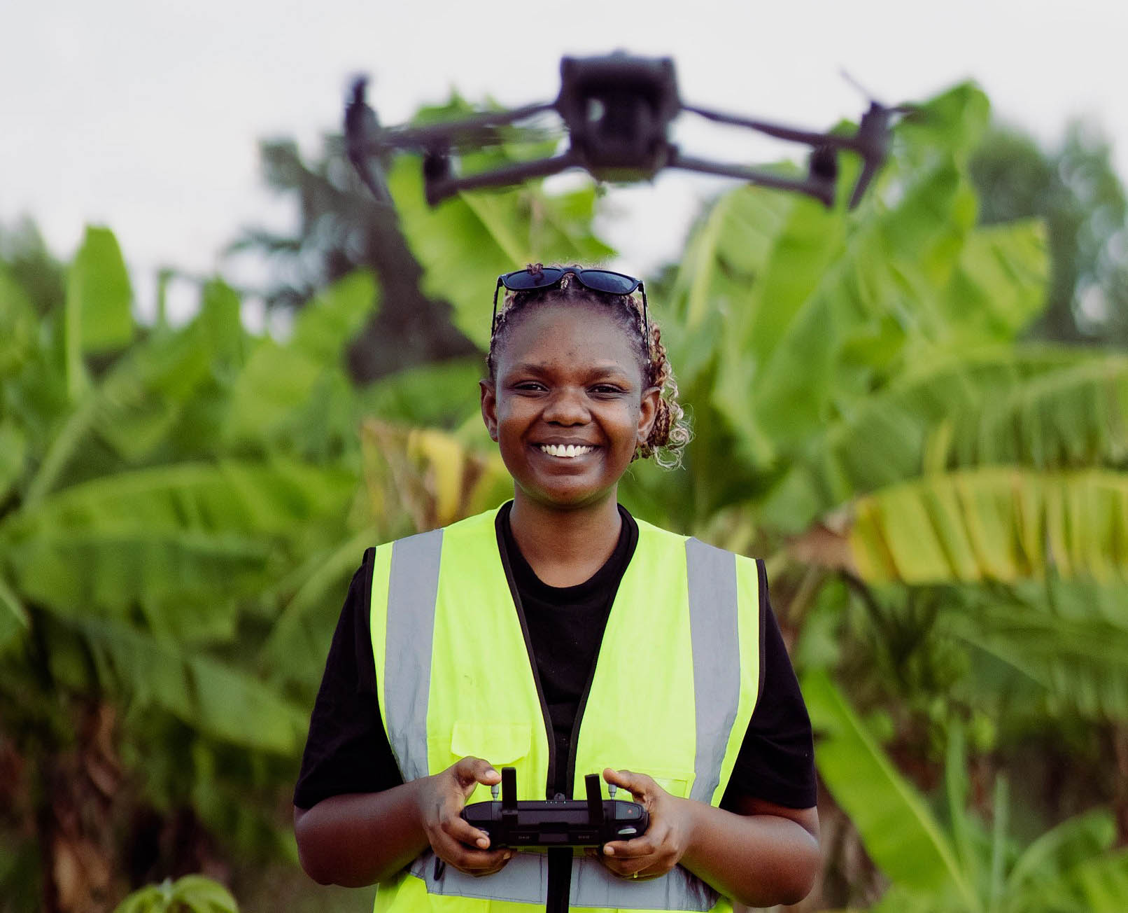 Rachel Kumwenda Kaunda wears yellow reflective safety vest and smiles as she flies a drone hovering above her head