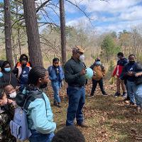 An agent stands in the forest with a group of children, teaching about natural resources.