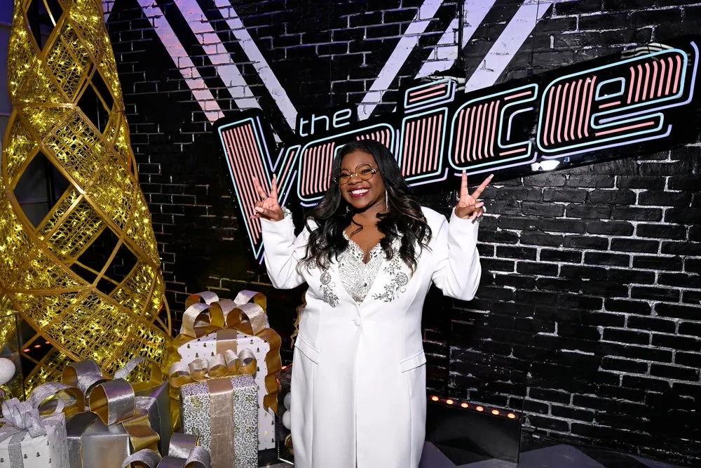 Jazz McKenzie flashes peace signs and smiles surrounded by Christmas gifts and "The Voice" sign behind her