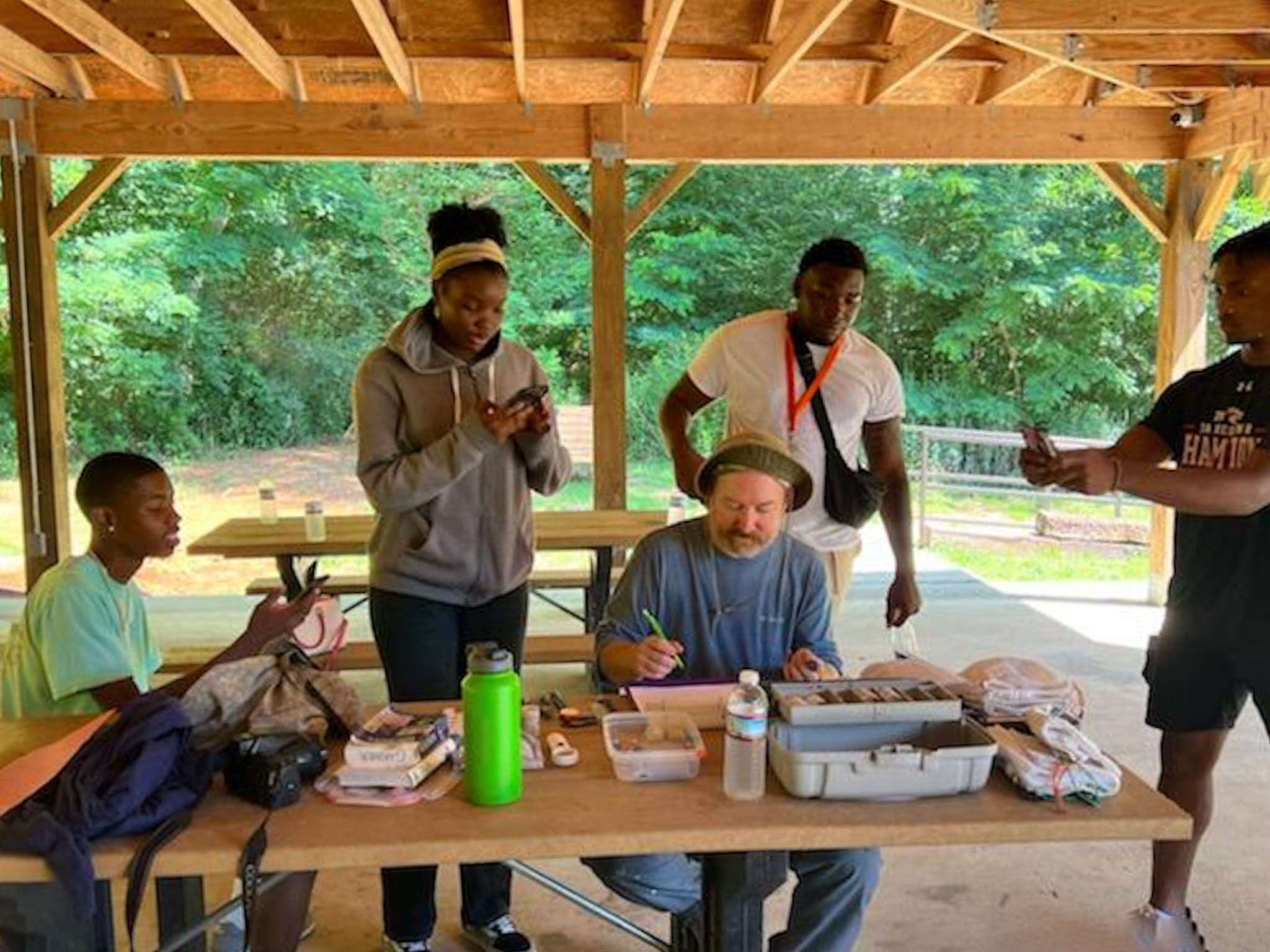 Thompson sits under a covered bench in the woods with four AAMU students working with him and standing around him