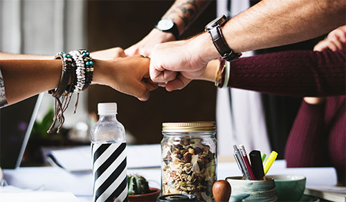 Five people fist-bump over a cluttered desk