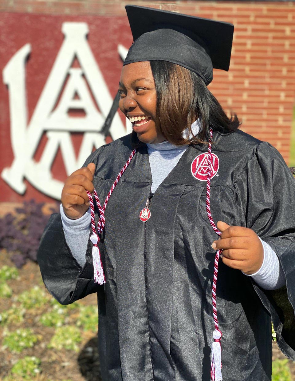 A girl smiling posing in graduation attire with giving cord.